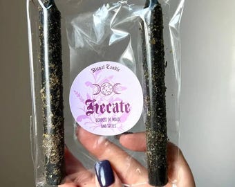 Puede incluir: Dos velas cónicas negras cubiertas de hierbas secas, empaquetadas en una funda de plástico transparente. Una etiqueta circular en el centro presenta el texto Ritual Candle, Hecate y Goddess of Magic and Spells, junto con un símbolo de triple luna.