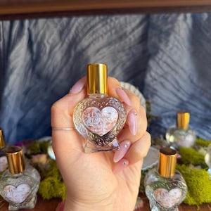 May include: A clear glass bottle with a gold cap and a heart-shaped design. The bottle is filled with a clear liquid and has the text "DIVINE FEMININE" and "PERFUME ROLLER" on the front.