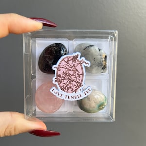 May include: A clear plastic box containing four tumbled stones: black, white, pink, and green. The box has a sticker that says "Love Tumble Set" with a pink background and a drawing of a cupid.