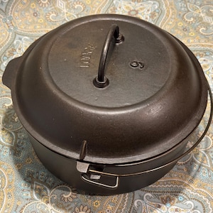 May include: A black cast iron Dutch oven with a lid and handle. The lid has the number 6 and the brand name "Lodge" embossed on it. The pot has a wire handle and a metal clasp.