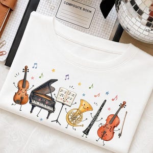 May include: A white t-shirt featuring a colorful graphic print of anthropomorphic musical instruments including a cello, a grand piano, a music stand with sheet music, a French horn, a clarinet, and a violin, all accented by small stars and notes.