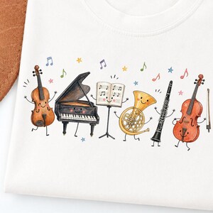 May include: A white t-shirt featuring a watercolor illustration of anthropomorphic musical instruments including a violin, a grand piano, a music stand with sheet music, a French horn, a clarinet, and a cello, all accented by colorful musical notes.