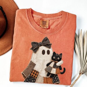May include: A burnt orange short-sleeve t-shirt featuring a fabric applique of a ghost wearing a polka dot bow and holding a black cat. The cat wears a small dress and bow. The ghost has a patchwork-style base with brown, black, and cream patterns.