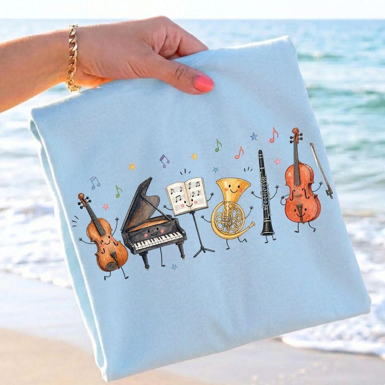 May include: A light blue sweatshirt featuring a colorful graphic print of anthropomorphic musical instruments including a violin, a grand piano, an open music book, a French horn, a clarinet, and a cello, all decorated with small floating musical notes.