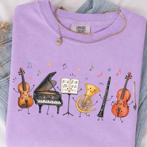 May include: A light purple t-shirt featuring a graphic of anthropomorphic musical instruments including a violin, a grand piano, a music stand with sheet music, a French horn, a clarinet, and a cello, all surrounded by colorful musical notes.