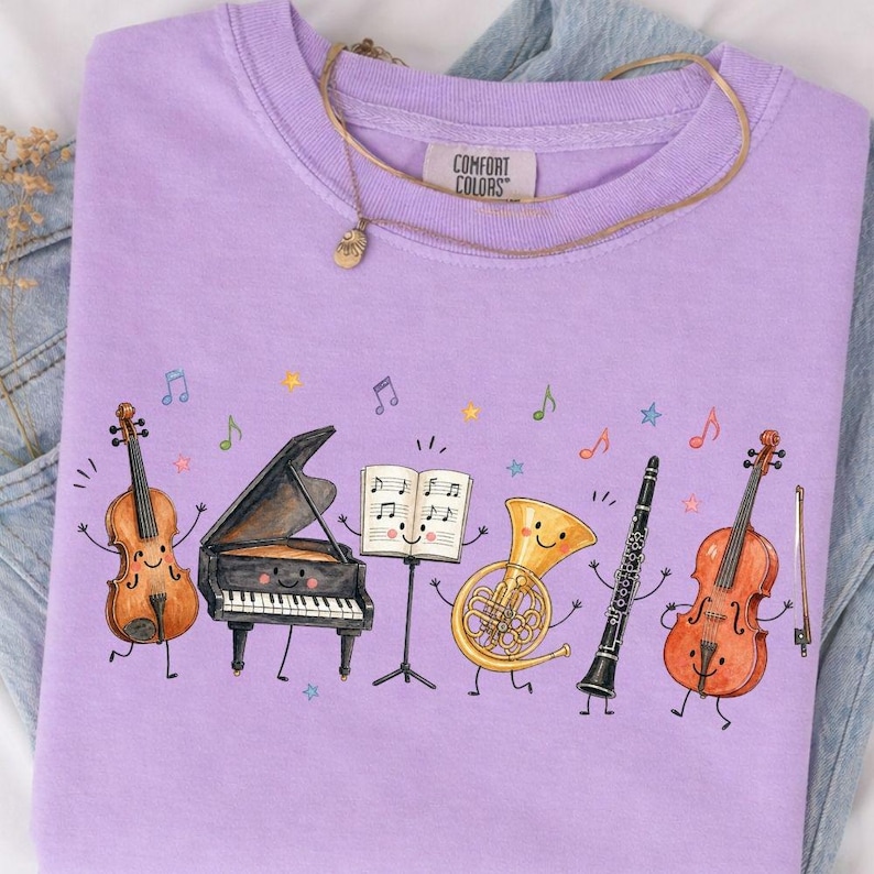 May include: A light purple t-shirt featuring a graphic of anthropomorphic musical instruments including a violin, a grand piano, a music stand with sheet music, a French horn, a clarinet, and a cello, all surrounded by colorful musical notes.