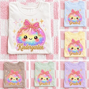 May include: A collection of t-shirts in various colours, each featuring a central graphic of a smiling, rainbow-coloured dumpling character with a pink glittery bow. The text below each character specifies grade levels including Kindergarten, Grade 1, Grade 2, Grade 3, Grade 4, and Pre-K.