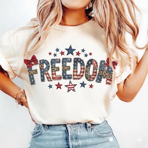 May include: Cream-colored t-shirt with the word "FREEDOM" in a patriotic design. The letters are a mix of red, white, and blue patterns, including stars, stripes, and leopard print. Red bows and stars surround the word, creating a festive look.