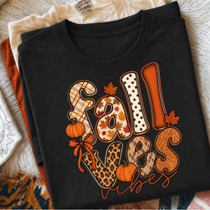 May include: A black crew neck t-shirt featuring the text fall vibes in large, stylised letters. The letters are filled with various patterns including plaid, polka dots, leopard print, and autumn leaves, accented by orange pumpkins and bows.