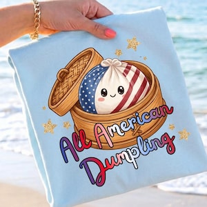 Könnte beinhalten: Hellblaues T-Shirt mit einem Cartoon-Knödel in einem Bambus-Dampfkorb. Der Knödel ist im Design der amerikanischen Flagge gehalten. Der Text "All American Dumpling" ist in einer bunten Schrift geschrieben. Goldene Glitzersterne sind verstreut.
