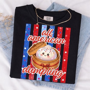 Könnte beinhalten: Schwarzes T-Shirt mit einem Aufdruck eines Knödels in einem Bambusdämpfer, mit dem Schriftzug "all american dumpling". Das Design beinhaltet rot-weiß-blaue Streifen und Sterne.