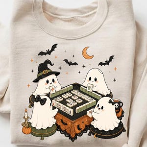 May include: A beige crewneck sweatshirt featuring a graphic of four white ghosts sitting around a square table playing mahjong. The illustration includes bats, a crescent moon, stars, a pumpkin, a candle, and a mug with a skull design.