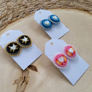 May include: Three pairs of beaded earrings on white tags. One pair is black and gold with a white heart, another is blue and white, and the third is pink and orange with a white heart. The earrings are on a wooden surface.