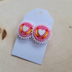 May include: Pink beaded heart-shaped earrings with white heart centers and white beaded edges. The earrings are displayed on a light blue tag against a light wood grain background. The earrings feature pink, yellow, and orange beads.