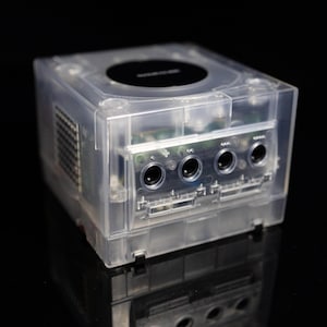 May include: A clear, translucent plastic video game console with a cube-shaped housing. The front panel features four circular controller ports and two memory card slots. A black circular disc cover is visible on the top surface of the console.