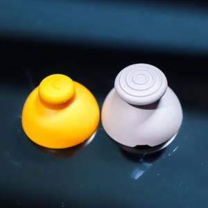 May include: Two small, round objects. One is yellow with a small, circular top. The other is light gray with a top featuring concentric circles. Both have a smooth, glossy finish and sit on a reflective surface.
