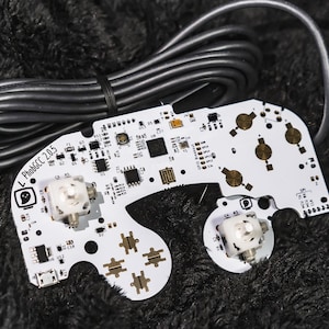 May include: A white circuit board with the text "PhobGCC 2.05" and a ghost logo. The board has various electronic components, including joysticks and buttons. A black cable is connected to the board.