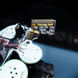 May include: Close-up of a disassembled circuit board with various components. The board features a white and black design with red and black wires, and a small rectangular section with yellow and black elements. The background is a dark, reflective surface.