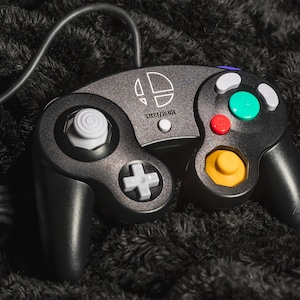 May include: Black GameCube controller with a Smash Bros. logo and Start/Pause text. Features a gray directional pad, a white analog stick, and colored buttons: green, red, and yellow. The controller has a wired connection.