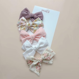 May include: Five assorted hair bows in shades of mauve, pink, and white, with floral and textured patterns. The bows are arranged on a white card with the brand name "mai & i" printed on it.