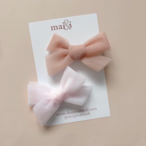 May include: Two tulle hair bows in pale pink and peach shades. The bows are displayed on a white card with the text "mai & i" and the website address www.shopmaiandi.com.