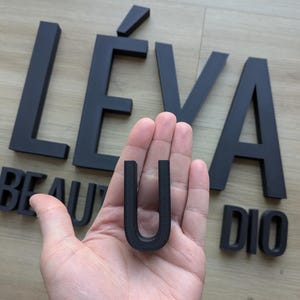May include: A set of matte black 3D-printed letters spelling out LÉVA BEAU U DIO, with the letter U held in the palm of a hand. The letters feature a clean, sans-serif font with a textured surface finish, arranged in a horizontal layout.