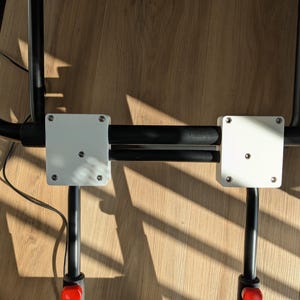 Op de afbeelding: Een zwart metalen frameconstructie met twee witte vierkante montageplaten die zijn vastgezet met zilverkleurige schroeven. De platen zijn bevestigd aan parallelle zwarte stangen, met extra zwarte buisvormige onderdelen en rode accenten.