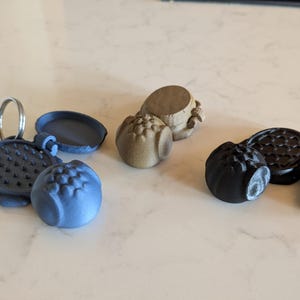 Könnte beinhalten: Eine Sammlung von 3D-gedruckten, kugelförmigen Objekten in verschiedenen Farben, darunter Blau, Schwarz und Beige. Einige haben strukturierte Oberflächen und aufklappbare Deckel, eines hat einen Schlüsselring. Auf hellem Untergrund.