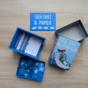 Könnte beinhalten: Brettspiel "Sea Salt & Paper" mit blauem und weißem nautischem Thema. Das Spiel enthält eine Box, Karten und blaue Spielsteine. Der Deckel der Box zeigt ein rotes und weißes Papierboot.