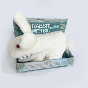 May include: A white plush rabbit toy with red eyes and visible teeth, presented in a decorative box. The box features the text "RABBIT WITH BIG" and a cage-like design. The rabbit has long ears and a fluffy tail.