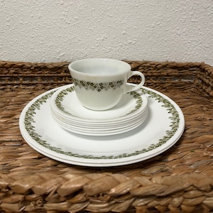 Könnte beinhalten: Ein Set aus weißen Keramiktellern und Tassen mit grünem Blumenmuster. Die Teller sind gestapelt und die Tasse steht oben auf dem Stapel.