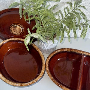 Pode incluir: Uma coleção de travessas de cerâmica marrom com acabamento brilhante e borda texturizada marrom claro. O conjunto inclui uma travessa oval, uma tigela redonda e uma travessa dividida. Um vaso de samambaia está ao fundo.