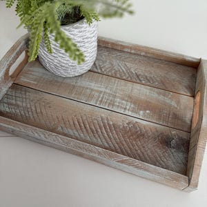 May include: A rectangular wooden serving tray with integrated handles, featuring a distressed, weathered finish in shades of brown and white. A small, textured, white ceramic pot with a green plant sits on the tray.