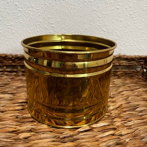 Puede incluir: Un macetero de metal dorado con un acabado martillado y una forma ligeramente redondeada. El macetero tiene una superficie lisa y brillante y un diseño simple y clásico.