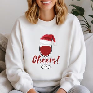 May include: A white long-sleeve sweatshirt featuring a graphic of a wine glass filled with red wine, topped with a red and white Santa hat. The word Cheers! is printed in red script lettering below the wine glass on the front of the garment.