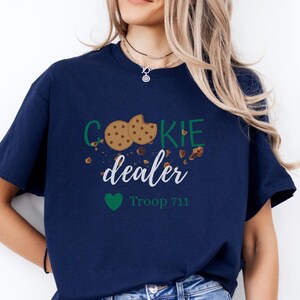 May include: Navy blue t-shirt with the words "COOKIE dealer" in green and white, featuring cookie graphics and the text "Troop 711" below a green heart. The shirt is a casual wear item.