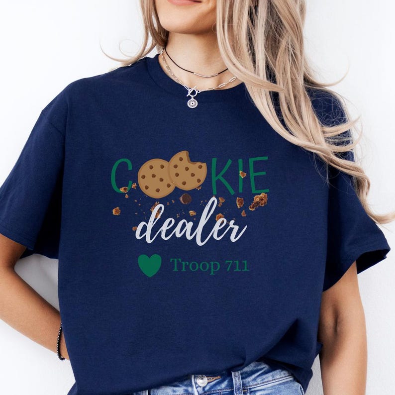 May include: Navy blue t-shirt with the words "COOKIE dealer" in green and white, featuring cookie graphics and the text "Troop 711" below a green heart. The shirt is a casual wear item.