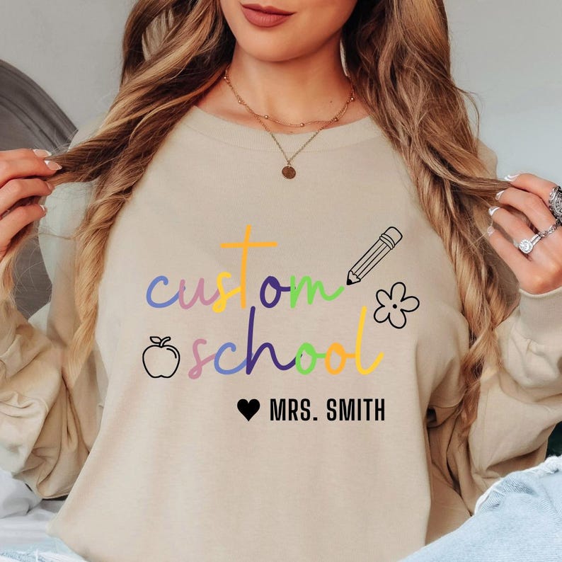Custom teacher sweatshirt with rainbow “custom school” lettering and personalized name text. Designed with apple, pencil, and flower icons, this unisex crewneck is a perfect mix of comfort and school pride, great for staff gifts, events, and classroom fashion.