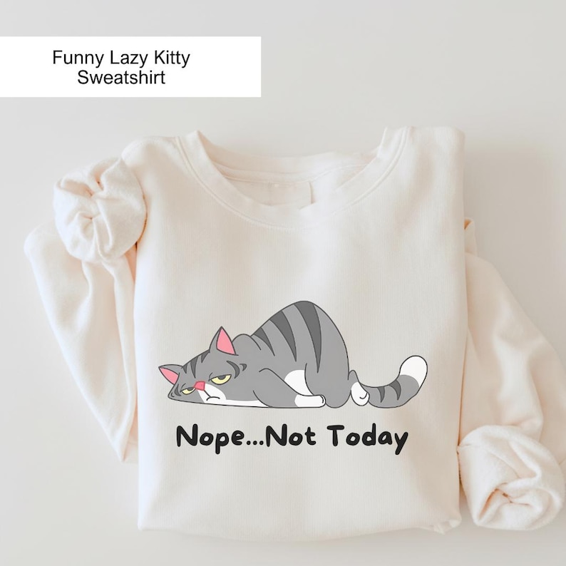 Funny cat sweatshirt featuring a grumpy cartoon cat lying down with the phrase “Nope… Not Today,” printed on a light gray cozy crewneck designed for cat lovers.