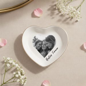 May include: A heart-shaped white ceramic trinket dish with a gold rim. The dish features a black and white photo of three people and the words "Besties Forever" in script.