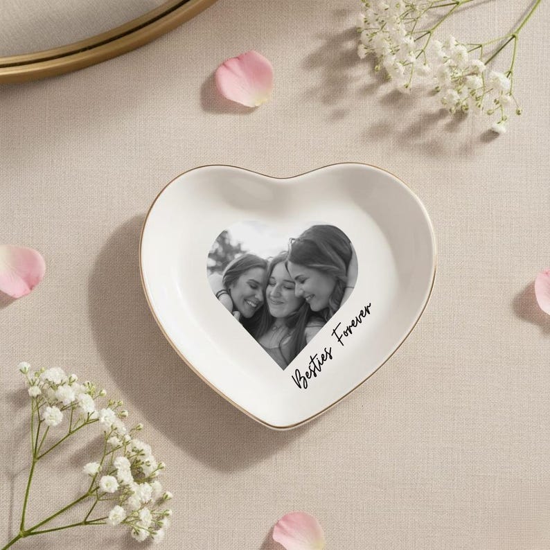 May include: A heart-shaped white ceramic trinket dish with a gold rim. The dish features a black and white photo of three people and the words "Besties Forever" in script.