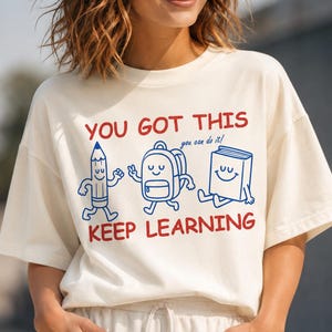 May include: An off-white short-sleeve t-shirt featuring red text that reads YOU GOT THIS and KEEP LEARNING. Between the text are blue line-art illustrations of a smiling pencil, a backpack, and a book, with the text you can do it! in blue.