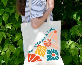 May include: Cream-colored tote bag with a floral design in orange, yellow, and teal. The words "ONE STEP AT A TIME" are printed on the bag. The bag is held by a person wearing a striped shirt, with a green leafy background.