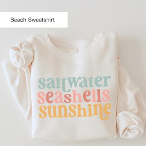 May include: Cream-colored sweatshirt with the words "saltwater seashells sunshine" in pastel colors. The text is stacked vertically. The sweatshirt has long sleeves and a crew neck. The words "Beach Sweatshirt" are in a white box at the top.