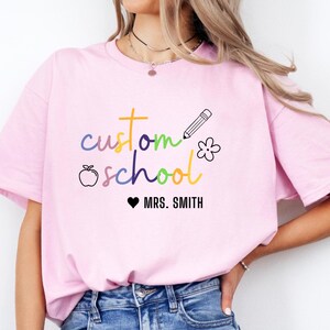 Personalized teacher sweatshirt with rainbow lettering “custom school” and teacher’s name in white text. Features playful icons like an apple, pencil, and flower. Perfect for showing school pride, this cozy crewneck makes a thoughtful teacher gift or staff spirit wear.