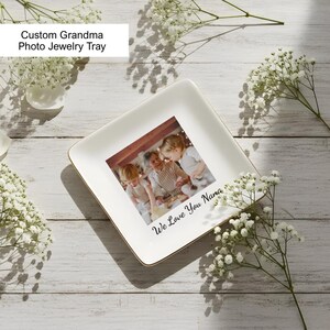 May include: A square white ceramic jewelry tray with a gold-toned rim. The center features a printed photograph of two children and an adult, with the black cursive text "We Love You Nana" printed below the image on the tray surface.