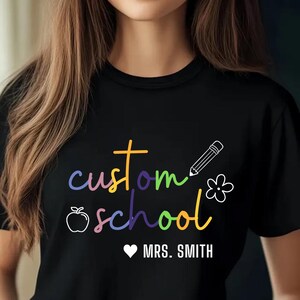 Cozy teacher crewneck sweatshirt customizable with school name and teacher’s name, featuring rainbow text and fun icons. A stylish and practical apparel piece for educators, making it a perfect gift for back-to-school, teacher appreciation, or everyday wear.