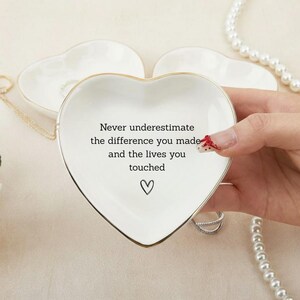 May include: White heart-shaped trinket dish with gold trim, featuring the text "Never underestimate the difference you made and the lives you touched" and a heart symbol. A small jewelry dish is visible in the background.