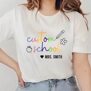 May include: White t-shirt featuring the words "custom school" in rainbow hues, a pencil, a flower, and an apple graphic. Below the text is "MRS. SMITH" with a heart symbol.