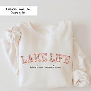 May include: Cream-colored sweatshirt with the words "LAKE LIFE" in a floral design. Below the text is the phrase "custom location" in a handwritten font. The top left corner has a white rectangle with the text "Custom Lake Life Sweatshirt".